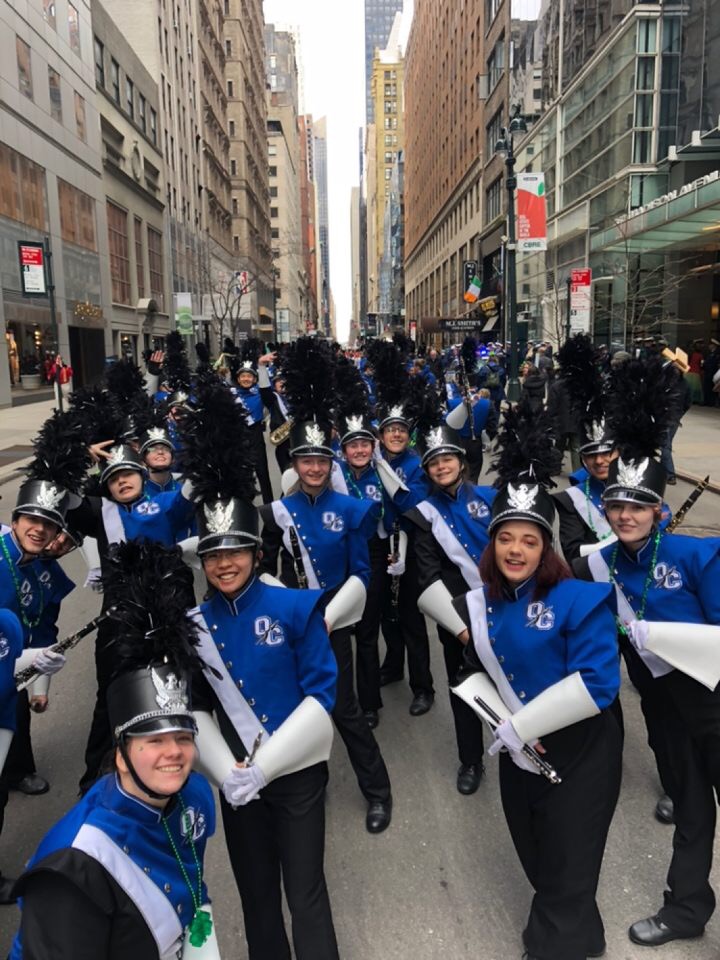 High school band members in blue uniforms gathered on the St. Patrick's Day Parade route